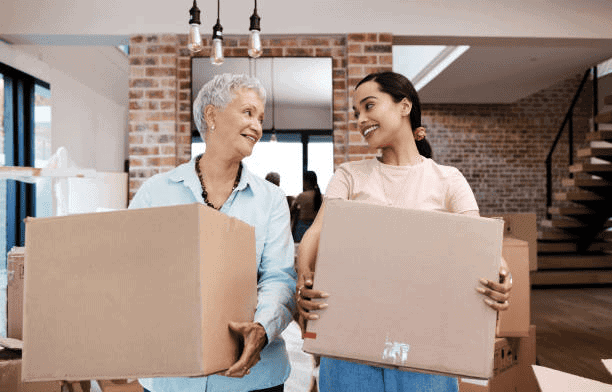 Two women smiling and carrying moving boxes in a modern home with brick walls and a staircase.