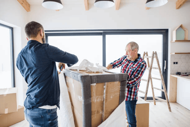 Two men one of them elderly unpacking a large cardboard box in a modern kitchen with large windows, wooden ceiling, and a ladder.