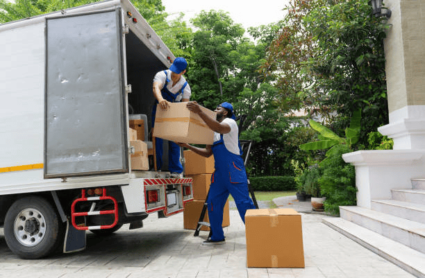 Two movers in blue uniforms loading cardboard boxes into a moving truck outside a house with lush greenery, symbolizing a relocation process.